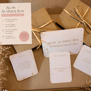 May include: A collection of items, including boxes and cards, with the text "SLOW SUMMER BOX" in a soft, neutral color palette. The boxes are tied with twine, and the cards feature floral accents and various prompts for summer activities.