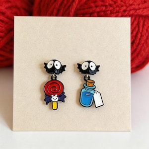 May include: A pair of enamel earrings with a black creature design and white eyes. One earring features a red and white swirled lollipop with a bat and a yellow stick. The other has a blue potion bottle with a cork and a white tag.