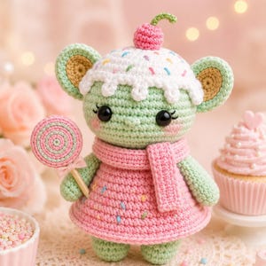 May include: A handmade crochet mouse doll wearing a pink dress and scarf, holding a lollipop. The mouse has a cupcake-shaped head with a cherry on top. The doll is green with pink cheeks and black eyes. A cupcake is in the background.