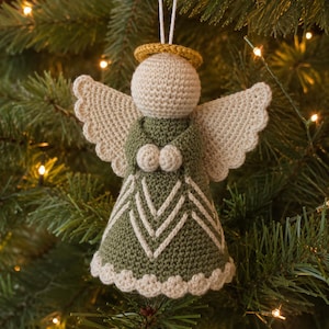 Op de afbeelding: Een gehaakte engel-ornament met een crèmekleurig hoofd en vleugels, een gouden halo en een olijfgroene jurk met witte chevron-details en een geschulpte witte zoom. Het figuurtje heeft kleine ronde handjes en een wit koord om op te hangen.