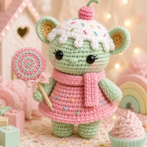 May include: A crocheted mouse doll with a cupcake-shaped head, wearing a pink dress and scarf, and holding a lollipop. The doll has a cherry on top and is decorated with colorful sprinkles. A cupcake and a rainbow are in the background.