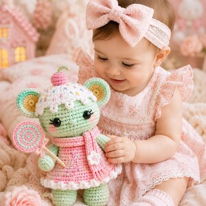 May include: A crochet doll in the shape of a mouse, wearing a pink dress and scarf, holding a lollipop. The doll has a green body, yellow ears, and a white hat with sprinkles. A baby in a pink dress and bow headband is holding the doll.