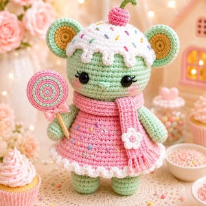 May include: A handmade crochet mouse doll with a cupcake-shaped hat, a pink dress, and a matching scarf. The doll holds a lollipop and has black eyes. The color palette includes pastel pink, green, white, and yellow.