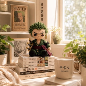 Può includere: Bambola di Zoro all'uncinetto della serie anime One Piece, con capelli e vestiti verdi e tre spade. La bambola è appoggiata su una pila di manga di One Piece. Sono visibili anche una tazza con testo giapponese e un poster "Wanted".