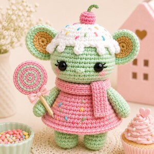 May include: A crocheted green creature wearing a pink dress and scarf, topped with a cupcake-shaped hat with a cherry. It holds a pink and green lollipop. The doll is on a white doily, with a cupcake and a bowl of sprinkles nearby.