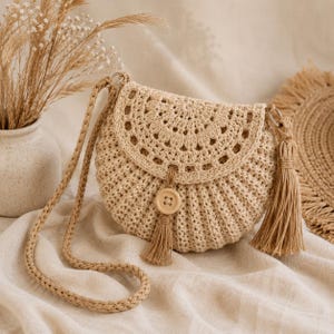 Peut inclure: Un sac à bandoulière en crochet beige de forme arrondie avec une fermeture à rabat. Le sac présente un motif floral décoratif sur le rabat, un bouton en bois et un pompon. La bandoulière est tressée.