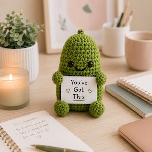 Può includere: Una statuetta di cetriolino lavorata all'uncinetto in verde con occhi a perla neri e un piccolo sorriso, che tiene in mano un cartello rettangolare bianco con la scritta "You've Got This" e due piccoli cuori verdi ai lati del testo.