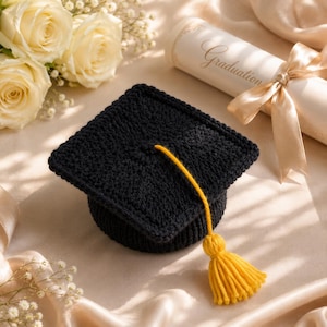 Könnte beinhalten: Eine schwarze, gestrickte Abschlusskappe mit gelber Quaste liegt auf einer satinartigen Oberfläche. Eine gerollte Urkunde mit Schleife und dem Wort "Graduation" ist zu sehen. Weiße Rosen und Schleierkraut befinden sich im Hintergrund.