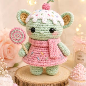 May include: A crocheted doll in the shape of a mouse, wearing a pink dress and scarf, and holding a lollipop. The mouse has a white and sprinkle-covered head, topped with a pink cherry. The doll is standing on a wooden slice.