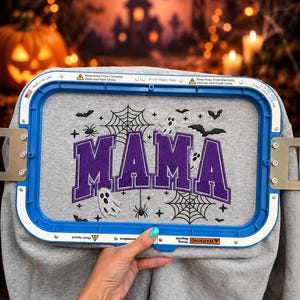 May include: A grey sweatshirt fabric held in a blue 8x13 inch embroidery hoop featuring the purple embroidered text MAMA surrounded by black spiderwebs, spiders, bats, and white ghost designs. The hoop frame includes safety warning text.