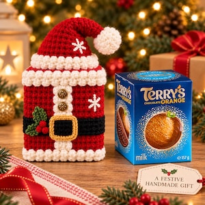 以下が含まれることがあります： Terry's Chocolate Orangeの箱用の赤と白のクロシェ編みサンタクロースカバー。カバーには、金色のバックルが付いた黒いベルト、2つの金色のボタン、赤い実が付いた緑のヒイラギの葉、白い雪の結晶の刺繍が施されています。