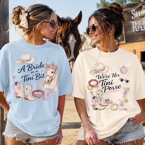 Könnte beinhalten: Ein hellblaues T-Shirt mit einer Grafik aus Cowboyhut, Pferd, Lasso und Martiniglas mit dem Text A Bride in a Tini Bit sowie ein cremefarbenes T-Shirt mit Cowboyhut, Lasso, Discokugel und dem Text We're Her Tini Posse.