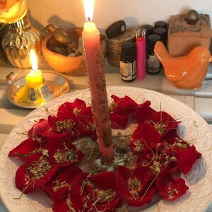 a white plate with a candle in the center, surrounded by red rose petals. The plate is placed on a white surface, and there are various objects in the background, including a mirror, a bird figurine, and a small vase.
