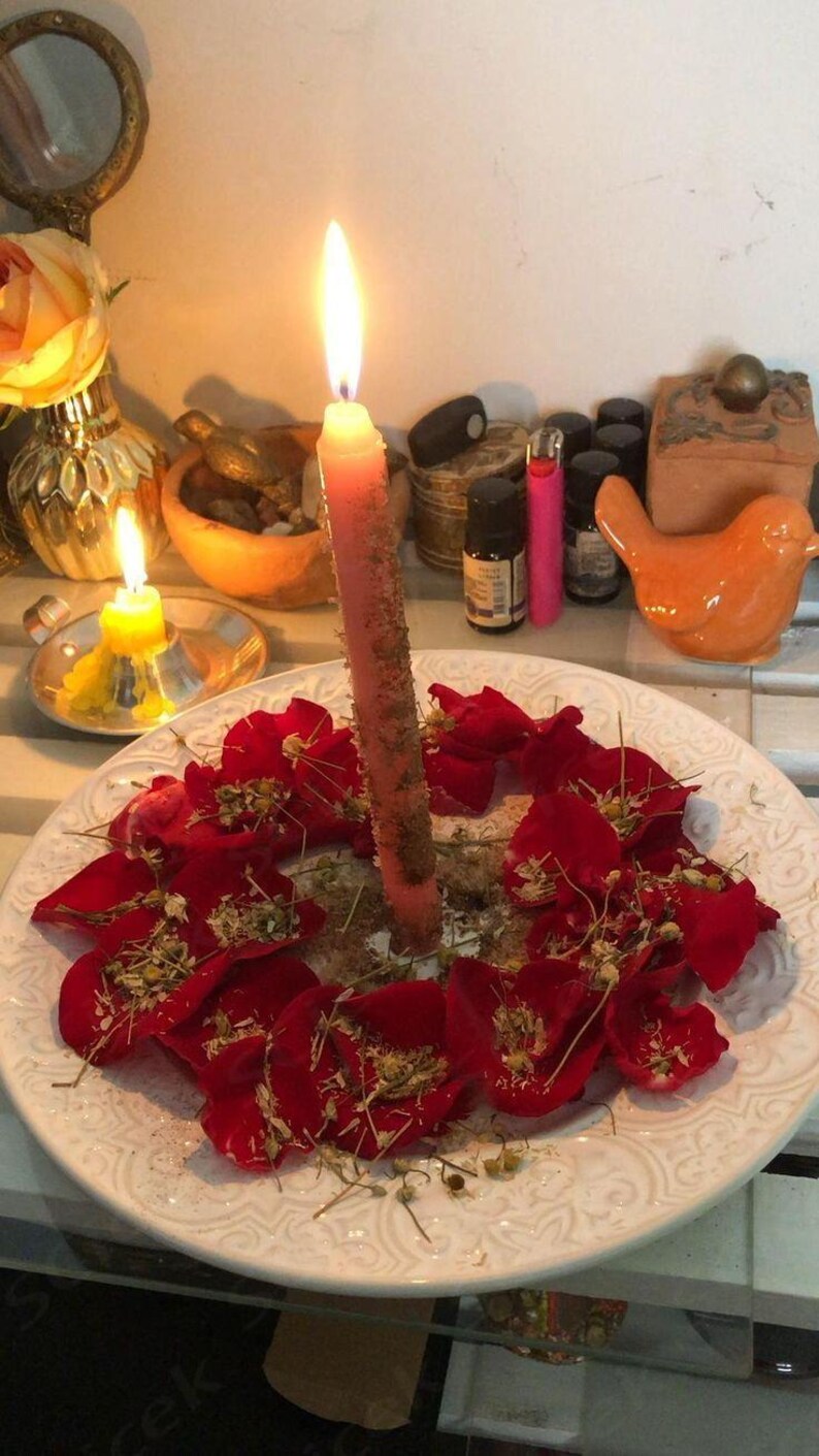 a white plate with a candle in the center, surrounded by red rose petals. The plate is placed on a white surface, and there are various objects in the background, including a mirror, a bird figurine, and a small vase.
