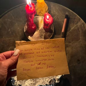 A hand holding a handwritten note with a candlelit candle and a pen on a table.