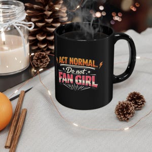 May include: A black ceramic mug with the text "ACT NORMAL.. Do not FAN GIRL" in a retro design. The mug is filled with a dark liquid, and steam rises from the top. The mug is on a light gray surface.