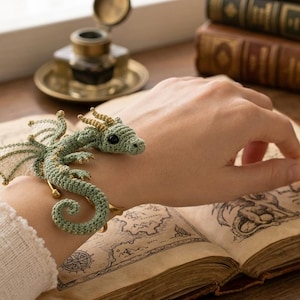 May include: A sage green crocheted dragon bracelet featuring intricate gold-toned thread detailing on the wings and horns, with a coiled tail and small black bead eyes, designed to wrap around the wrist as a decorative accessory.