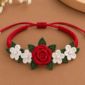 May include: A red adjustable cord bracelet featuring a central crocheted red rose surrounded by smaller white crocheted flowers and dark green crocheted leaves. The ends of the red cord are finished with small white beads and red knotted spheres.
