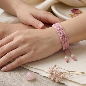 May include: A light pink crochet bracelet featuring a textured woven pattern and adjustable drawstring closure. The ends of the bracelet are finished with two small white pearl beads and matching pink thread knots, creating a delicate accessory.