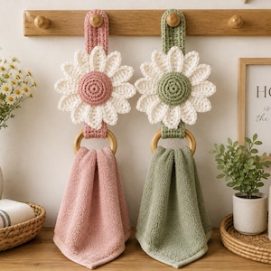 May include: Two hanging hand towels featuring crocheted daisy toppers in pink and sage green. Each towel is suspended from a wooden ring attached to a matching crocheted strap. The daisy centers match the towel colors, with white petals.