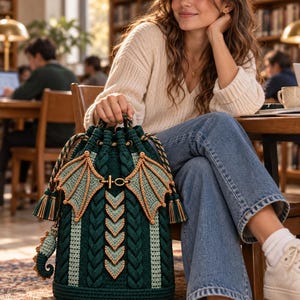 Puede incluir: Un bolso tipo cubo de ganchillo en color verde oscuro con un patrón de punto trenzado texturizado, detalles de alas de dragón en verde claro, un cierre central de palanca y un dije de cola de dragón con borlas a juego.