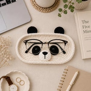 May include: A cream-colored, crocheted panda-shaped tray holding eyeglasses. The tray has black crocheted ears and eyes, and a white and black nose. A pair of black-framed glasses rests inside the tray. The tray is on a neutral-colored surface, with a watch and rings.