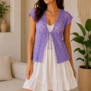 Puede incluir: Un chaleco de ganchillo color lavanda con un intrincado patrón floral, mangas cortas y un delicado cierre de lazo frontal, sobre un sencillo vestido blanco sin mangas con un dobladillo con volantes que cae sobre la rodilla.