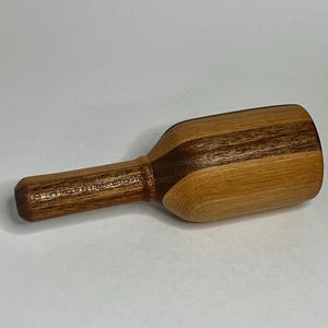 May include: A wooden scoop featuring a cylindrical handle and a wide, rounded bowl. The object is crafted from two distinct types of wood, showcasing a dark brown grain on the handle and a lighter, honey-toned wood on the bowl section.