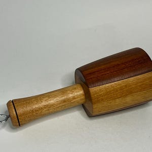 May include: A wooden mallet featuring a cylindrical handle made of light-toned wood and a head constructed from contrasting segments of light and dark brown wood. A small metal eye bolt is attached to the end of the handle for hanging.