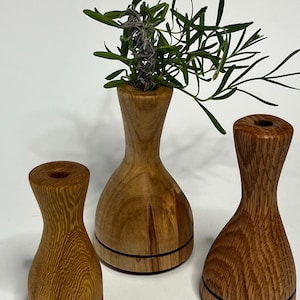 May include: Three wooden bud vases with a natural finish. One vase holds green and purple sprigs. The vases have a bulbous base and a narrow neck. Each vase has a black line around the base.