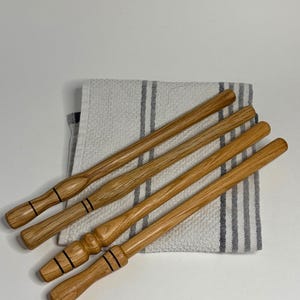 Puede incluir: Cuatro palos de madera largos y delgados con un acabado de grano de madera marrón claro natural. Cada palo presenta mangos torneados decorativos con finos anillos negros horizontales. Los palos están dispuestos en diagonal sobre un paño a rayas blancas y grises.