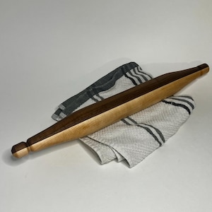 May include: A long, cylindrical rolling pin crafted from two distinct types of wood, featuring a dark brown walnut section and a lighter tan maple section, resting diagonally across a folded white and grey striped kitchen towel.