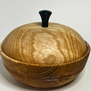 Könnte beinhalten: Eine Holzschale mit Deckel. Die Schale besteht aus hellem Holz mit einem natürlichen Maserungsmuster. Der Deckel hat einen schwarzen Knopf oben. Die Schale hat eine runde Form und eignet sich zur Aufbewahrung kleiner Gegenstände.