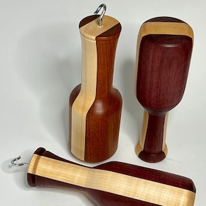 May include: Three handcrafted wooden objects with smooth, polished surfaces. Two objects are upright and one lies horizontally. They feature contrasting light cream and deep reddish-brown wood segments joined together. Two include a small metal hook.