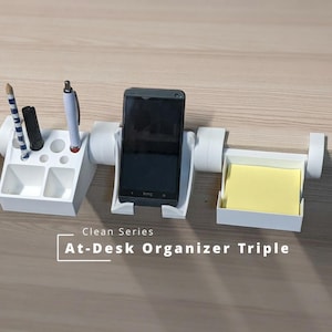 Peut inclure: Un organisateur de bureau en plastique blanc avec trois sections fixées au bord d'un bureau. La section gauche contient des stylos, le centre un smartphone et la droite des notes adhésives jaunes. Texte : Clean Series At-Desk Organizer Triple.