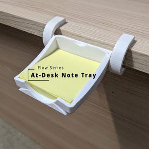 May include: A white plastic note tray with two curved clips that attach to the edge of a desk. The tray holds a stack of square yellow sticky notes. The text Flow Series At-Desk Note Tray is printed in black on the surface of the note stack.