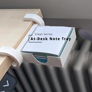 Könnte beinhalten: Eine weiße Notizablage aus Kunststoff mit einem Klemmmechanismus, der an der Schreibtischkante befestigt ist. Die Ablage enthält einen Stapel quadratischer Notizzettel. Der Text Clean Series At-Desk Note Tray ist in Schwarz aufgedruckt.