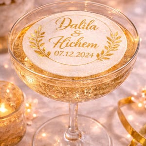 May include: A clear glass coupe filled with a golden liquid, featuring a round white topper printed with the gold text Dalila & Hichem and the date 07.12.2024, framed by two gold laurel leaf branches on a circular border.
