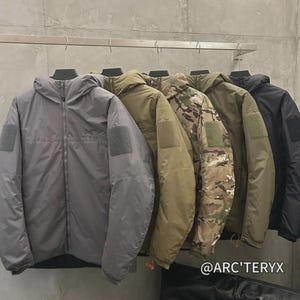 May include: Several hooded jackets in shades of grey, olive green, camouflage, and black, suspended from a metal rail. Each jacket features a sleeve patch and a zip fastening. The brand name "@ARC'TERYX" is displayed.