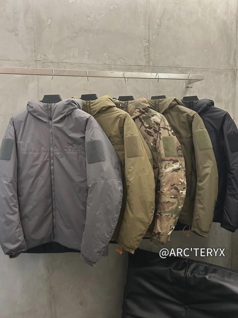 May include: Several hooded jackets in shades of grey, olive green, camouflage, and black, suspended from a metal rail. Each jacket features a sleeve patch and a zip fastening. The brand name "@ARC'TERYX" is displayed.