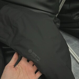 May include: A black jacket with the GORE-TEX INFINIUM logo. The jacket appears to be made of a smooth, matte material. The image shows a close-up of the jacket's sleeve.