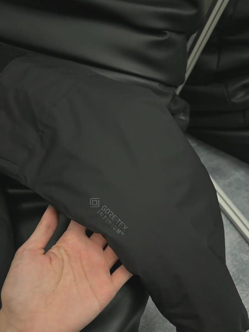 May include: A black jacket with the GORE-TEX INFINIUM logo. The jacket appears to be made of a smooth, matte material. The image shows a close-up of the jacket's sleeve.