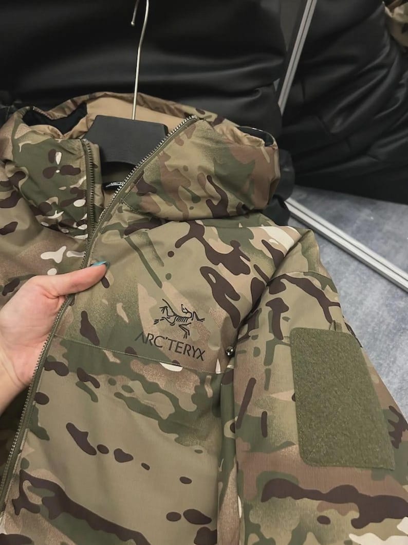 May include: Camouflage jacket with the Arc'teryx logo. The jacket has a zip, a hood, and a green patch on the sleeve. The colour scheme includes shades of green, brown, and tan.
