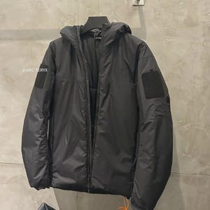 May include: Black hooded jacket with a zip fastening, hanging on a rail. The jacket features a patch on the left sleeve and the brand name is visible on the left side. The jacket has a drawstring at the bottom.