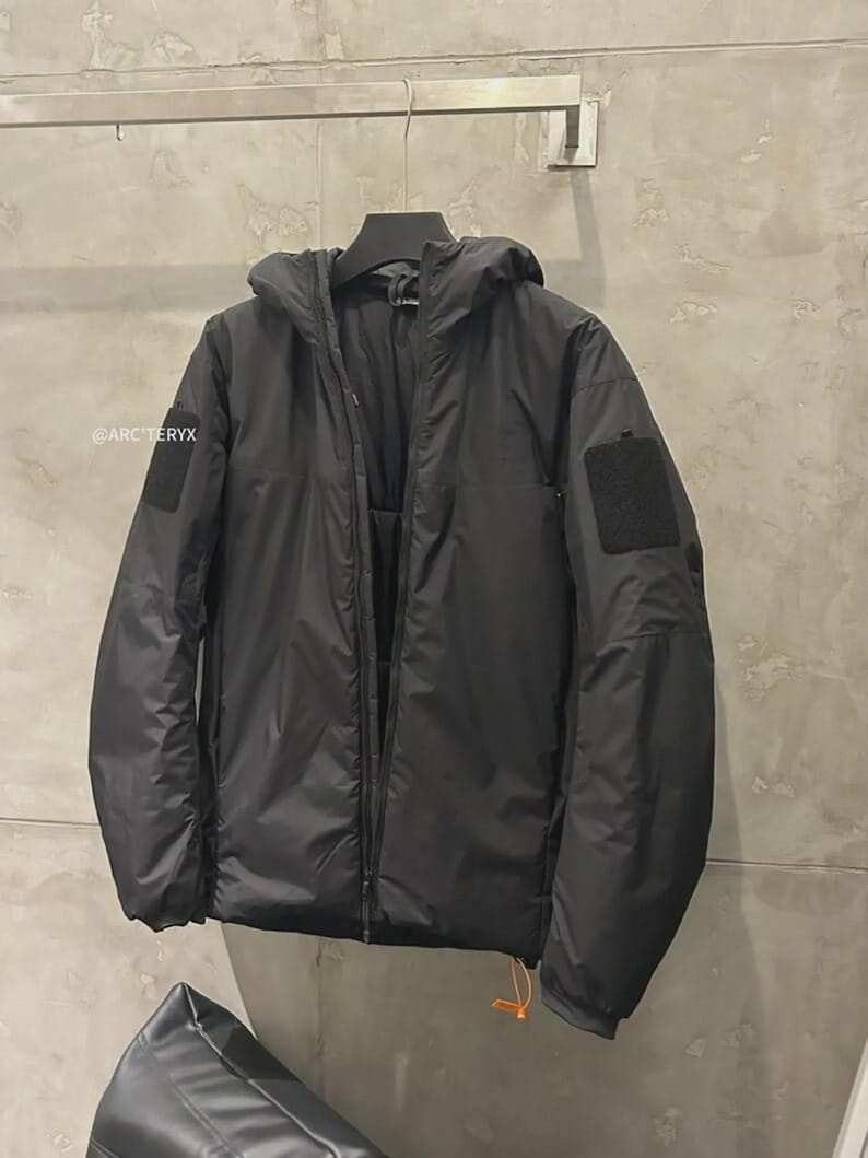 May include: Black hooded jacket with a zip fastening, hanging on a rail. The jacket features a patch on the left sleeve and the brand name is visible on the left side. The jacket has a drawstring at the bottom.