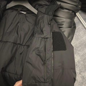 May include: Black puffer jacket and vest set. The jacket has a hood and is hanging on a hanger. The vest has a zip closure and a small pocket. The vest also has a black patch.