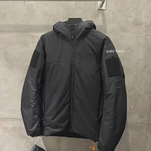 May include: Black hooded jacket with a full-length zip, two square patches on the sleeves, and a small orange pull tab. The jacket has a minimalist design and is hanging on a metal bar. The brand name "@ARC'TERYX" is visible on the right sleeve.