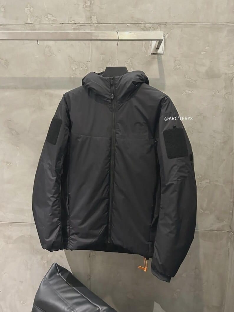 May include: Black hooded jacket with a full-length zip, two square patches on the sleeves, and a small orange pull tab. The jacket has a minimalist design and is hanging on a metal bar. The brand name "@ARC'TERYX" is visible on the right sleeve.