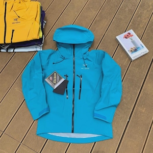 May include: A turquoise hooded jacket with a black zipper and two front pockets. The jacket has the brand name "Arc'teryx" on the chest and a "GORE-TEX" tag. Other jackets in yellow, purple, and white are stacked in the background.