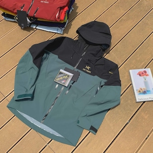 May include: A teal and black hooded jacket with a full-length zipper. The jacket has the brand name "Arc'teryx" on the chest and a "GORE-TEX" tag. Several other jackets are stacked in the background.